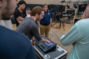 Instructor demonstrating a digital audio mixing console to a group of students in a live sound or production lab, with stage lighting and equipment visible in the background.