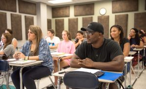 Students sitting in desks, with notebooks open, focused toward the front of the classroom.