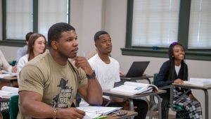 Students sitting at desks in classroom, with books and laptops open.
