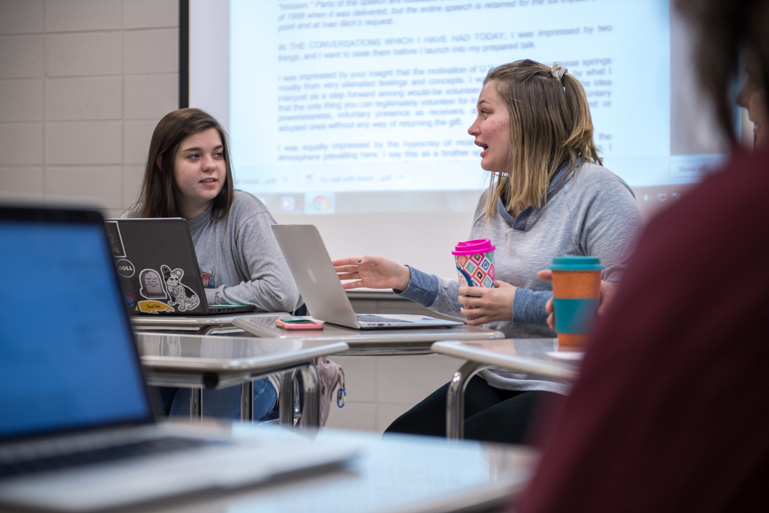 Two students in class with laptops, sitting at desks and discussing assignment.