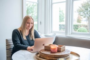 Woman sitting at a round table by a window, working on a laptop with a tray of candles and woven containers in the foreground.