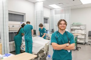 Nursing student wearing scrubs and a stethoscope stands in a clinical stimulation lab while other students practice patient care in the background.