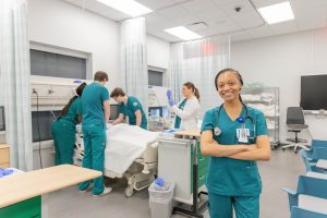 Nursing student wearing scrubs and a stethoscope stands in a clinical stimulation lab while classmates practice patient care in the background.
