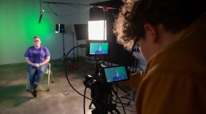 Student sitting in a chair with video equipment and lighting placed around her, while another student operates a camera on a tripod with visible displays.