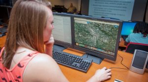 Student using GIS software to analyze map data on dual computer monitors.