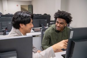 Professor sitting next to student in a computer lab, while pointing at students computer screen.