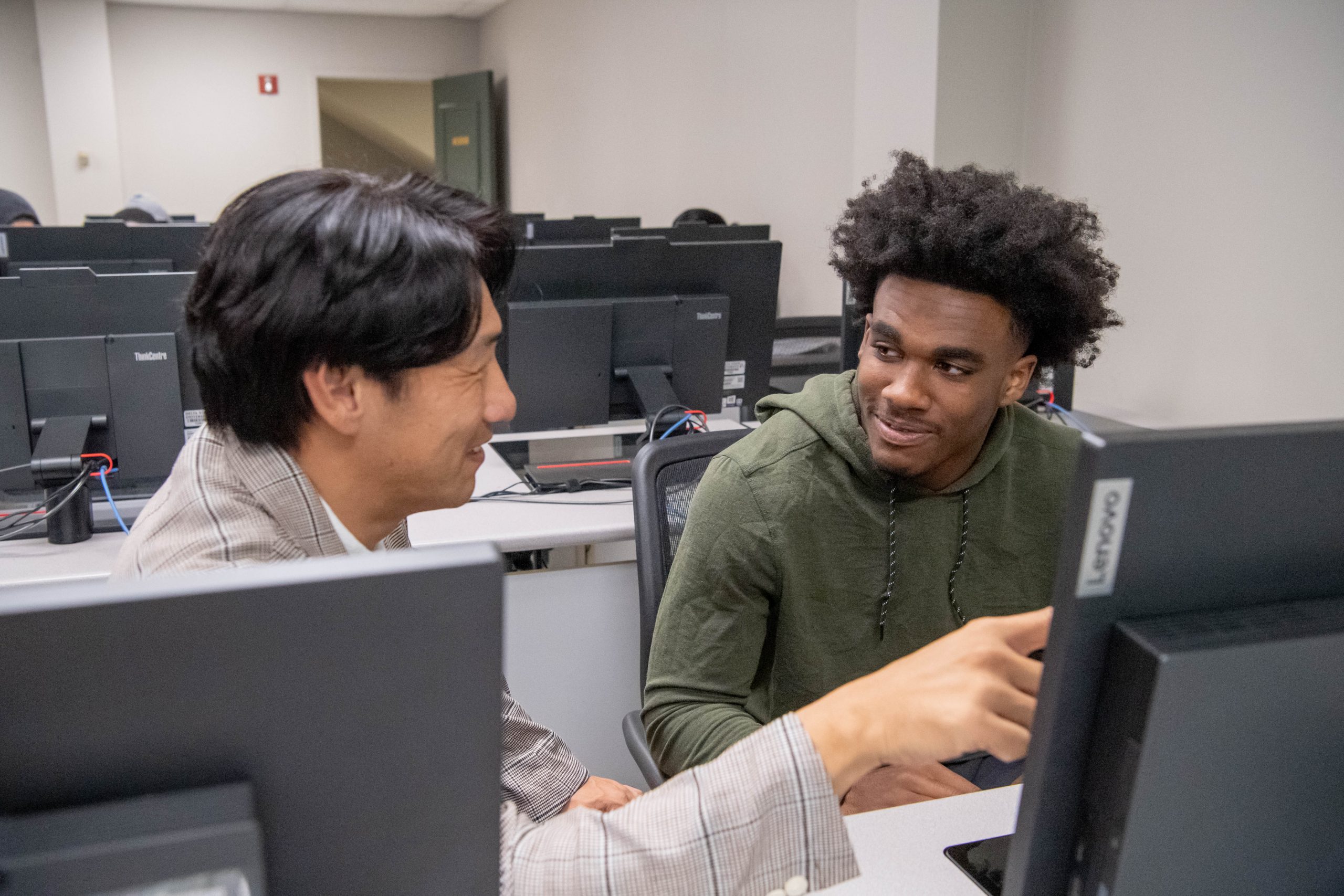 Professor sitting next to student in a computer lab, while pointing at students computer screen.