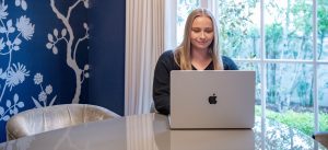 Student sitting at dining room table, with laptop and large window behind her at home.