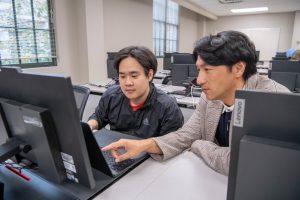 Professor sitting next to student in a computer lab, while pointing at students computer screen.