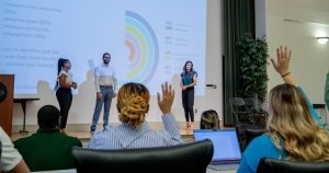 Three students answering questions from people with hands raised in the audience, about data on large PowerPoint displayed behind them.