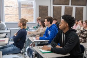 Students sitting at desks in a classroom, attentively listening, with one student in the foreground wearing a black hoodie.