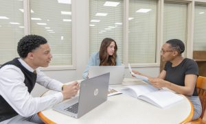 Three students sitting at a table with books and a laptop discussing a mock case.
