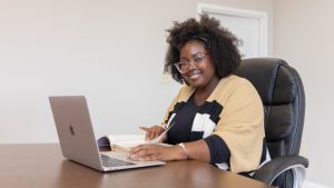 A woman sitting at a table in a school conference room, smiling with a computer in front of her.