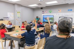 An elementary classroom filled with students sitting at desks, some raising a hand along with their teacher, as an administrator observes.