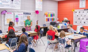 A teacher standing near a board in front of elementary students in a classroom with one student raising their hand.