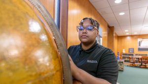 A female student wearing a Delta State University polo shirt and glasses closely examining a large globe in a well-lit library study space with bookshelves and seating areas.