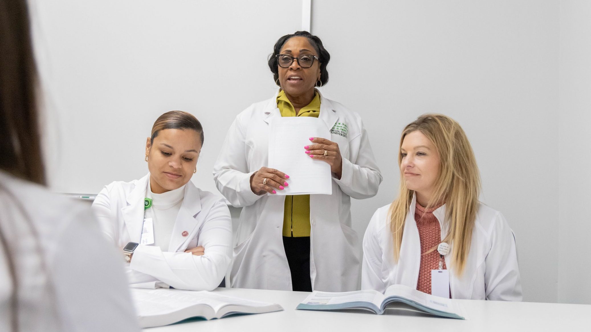 Three women in lab coats, one standing and speaking while the other two sit with open books.
