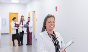 Smiling woman in a lab coat holding a clipboard in the foreground, with three other women in lab coats talking in the hallway behind her.
