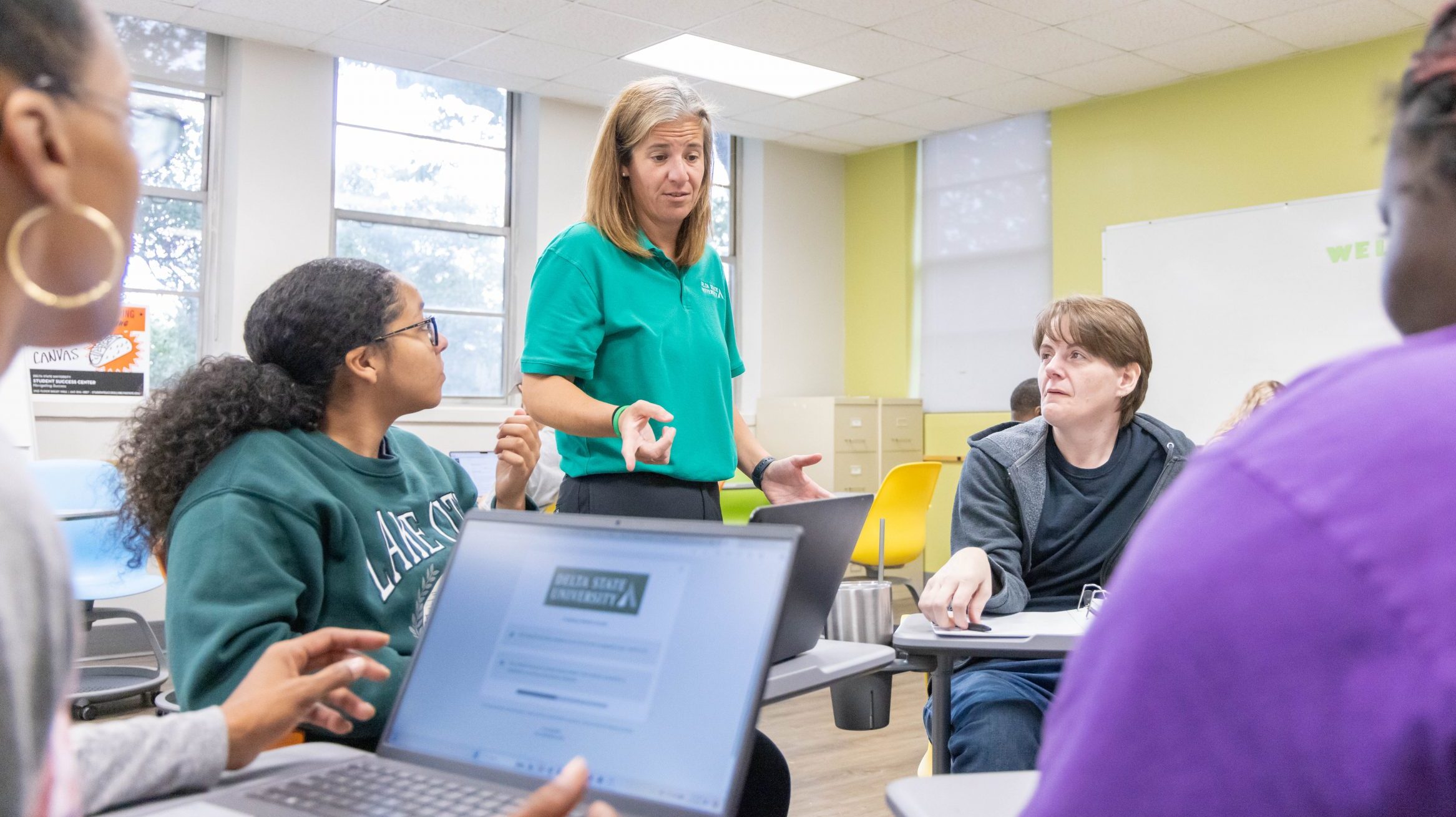 Instructor helping a group of students sitting at desks organized in a circle. Each student is looking at the instructor.