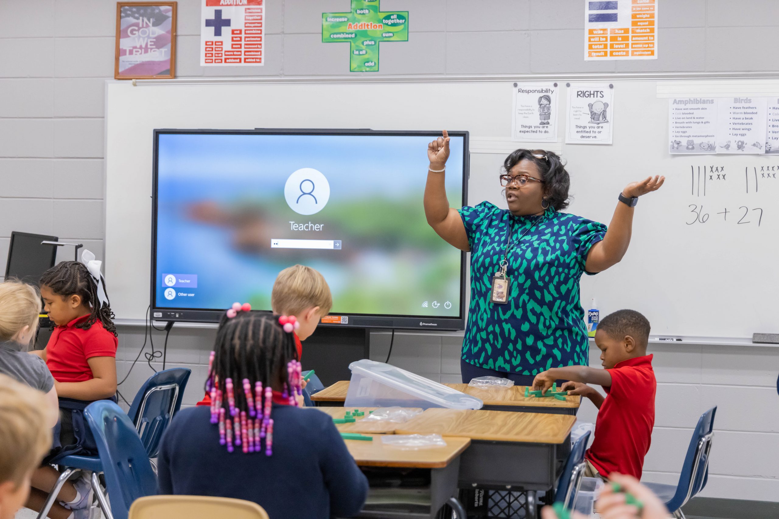 A teacher standing in front of elementary students.in a classroom with arms raised and students conducting activity at desks.