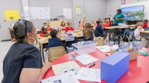 A school administrator sitting at a table at the back of a classroom, observing a teacher teaching elementary students.