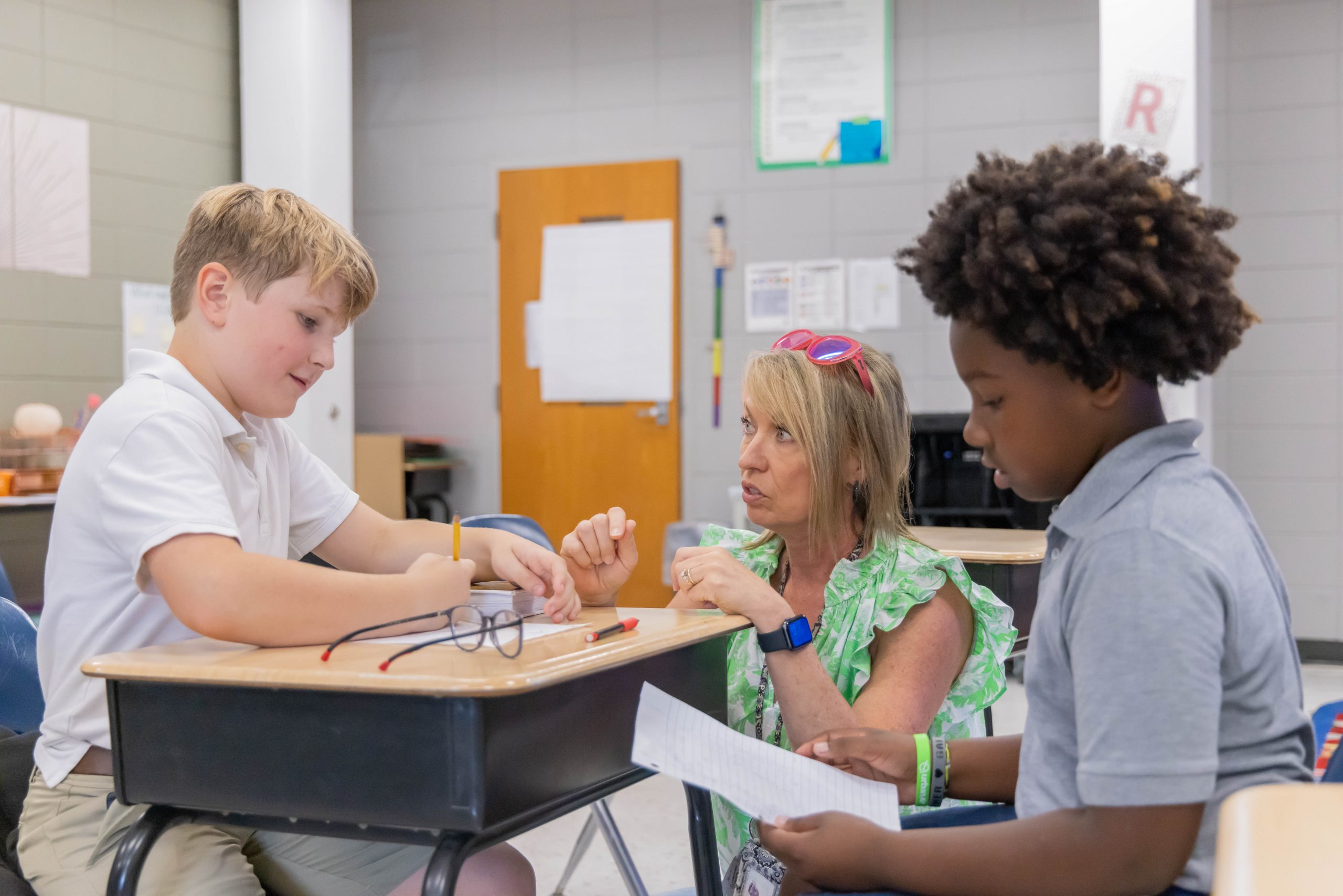 Two student grouped together at a desk with a teacher crouched down at the desk to help them with their class assignment.