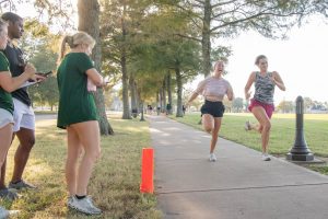 Two runners sprinting on a walking trail surrounded by trees and grassy areas, with three individuals standing nearby observing and recording the activity.