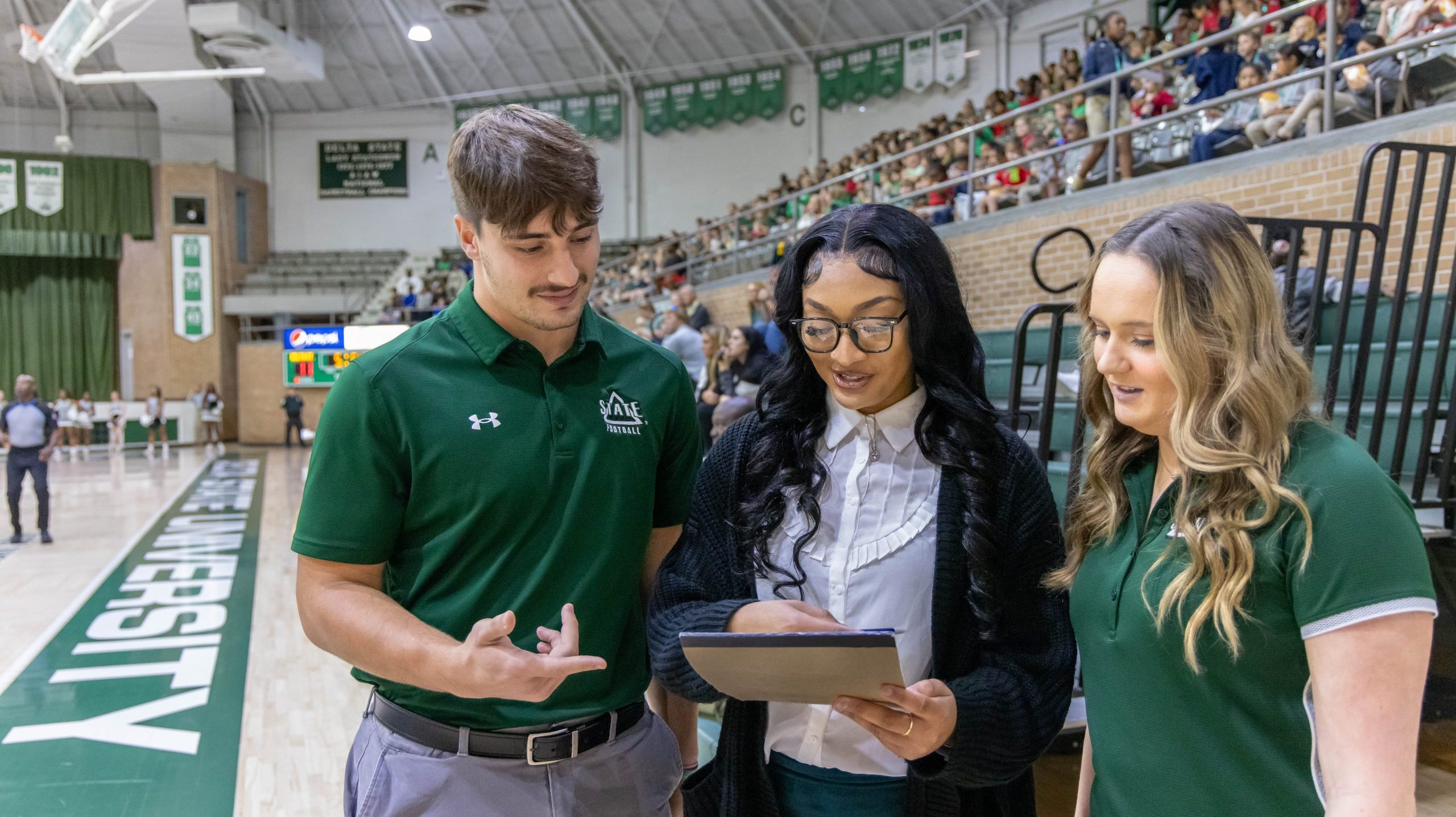 Three students standing inside a gymnasium engaged in a discussion around information on a tablet during a basketball game.