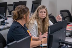 Two students sitting at computer desks in a classroom, talking and collaborating on an assignment.