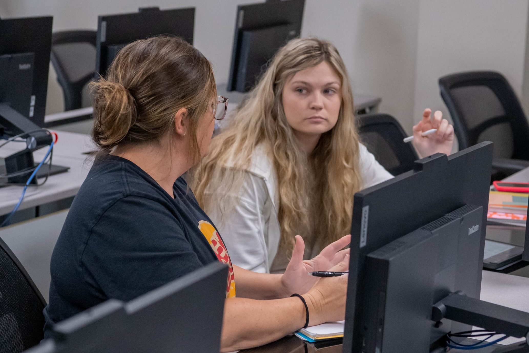 Two students sitting at computer desks in a classroom, talking and collaborating on an assignment.