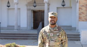 U.S. Army service member in uniform standing in front of a campus building with arched entryways.