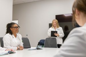 Three MSN students in lab coats engaging in a discussion during a meeting, one seated and the other standing with a tablet.