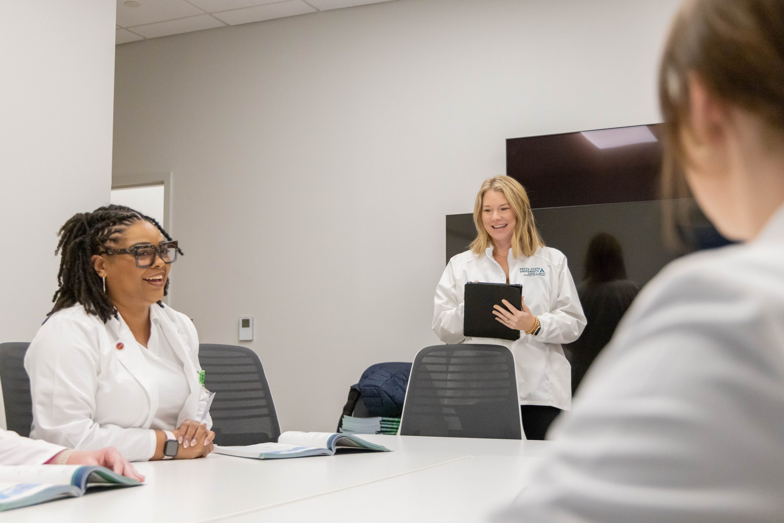 Three women in lab coats in a discussion during a meeting, two seated and the other standing with a tablet.