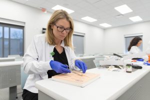 Woman in a lab coat and gloves practices suturing on a training pad in a clinical classroom setting.
