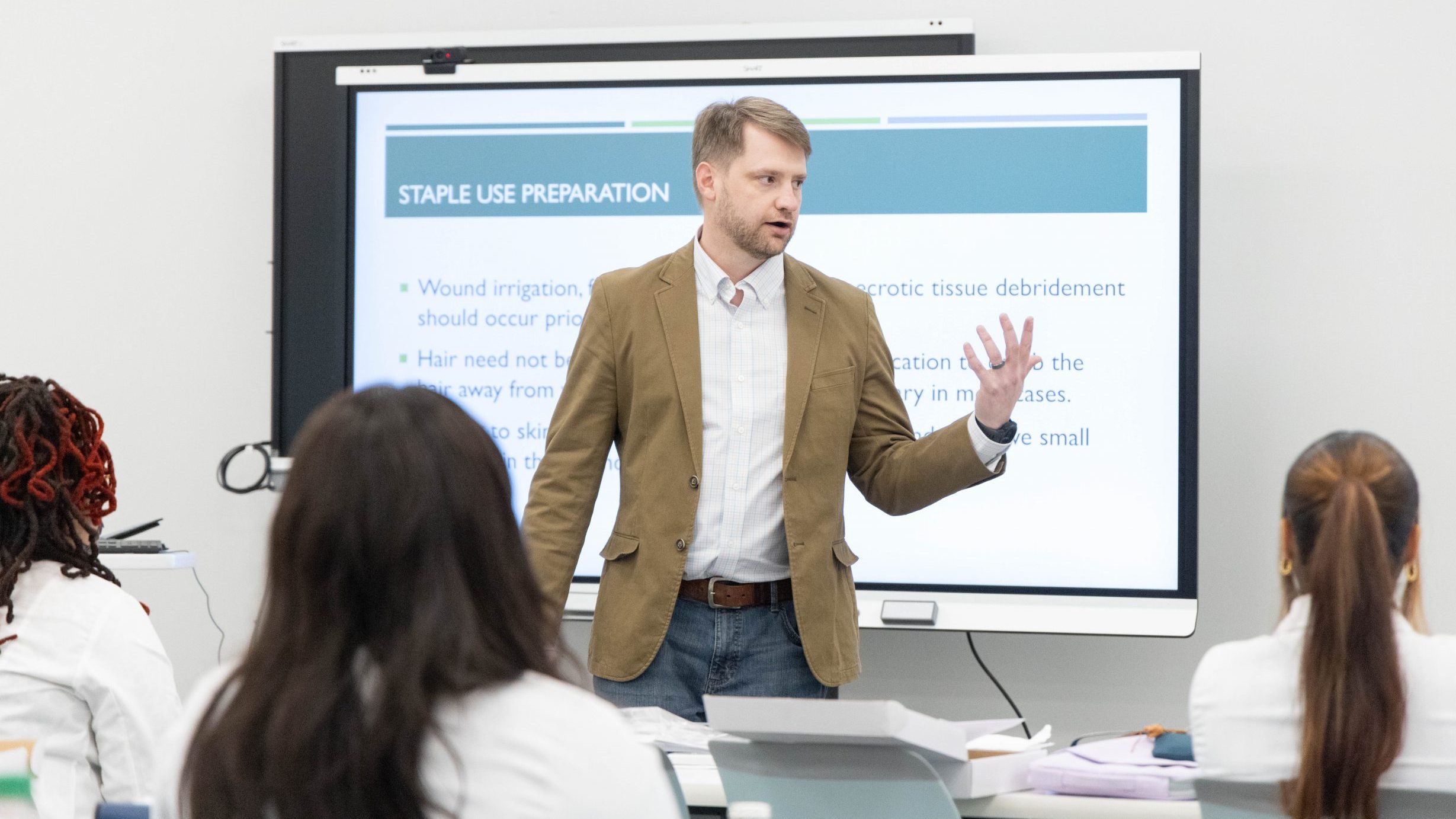 A male professor stands in front of a screen giving a presentation to a group of people in lab coats.