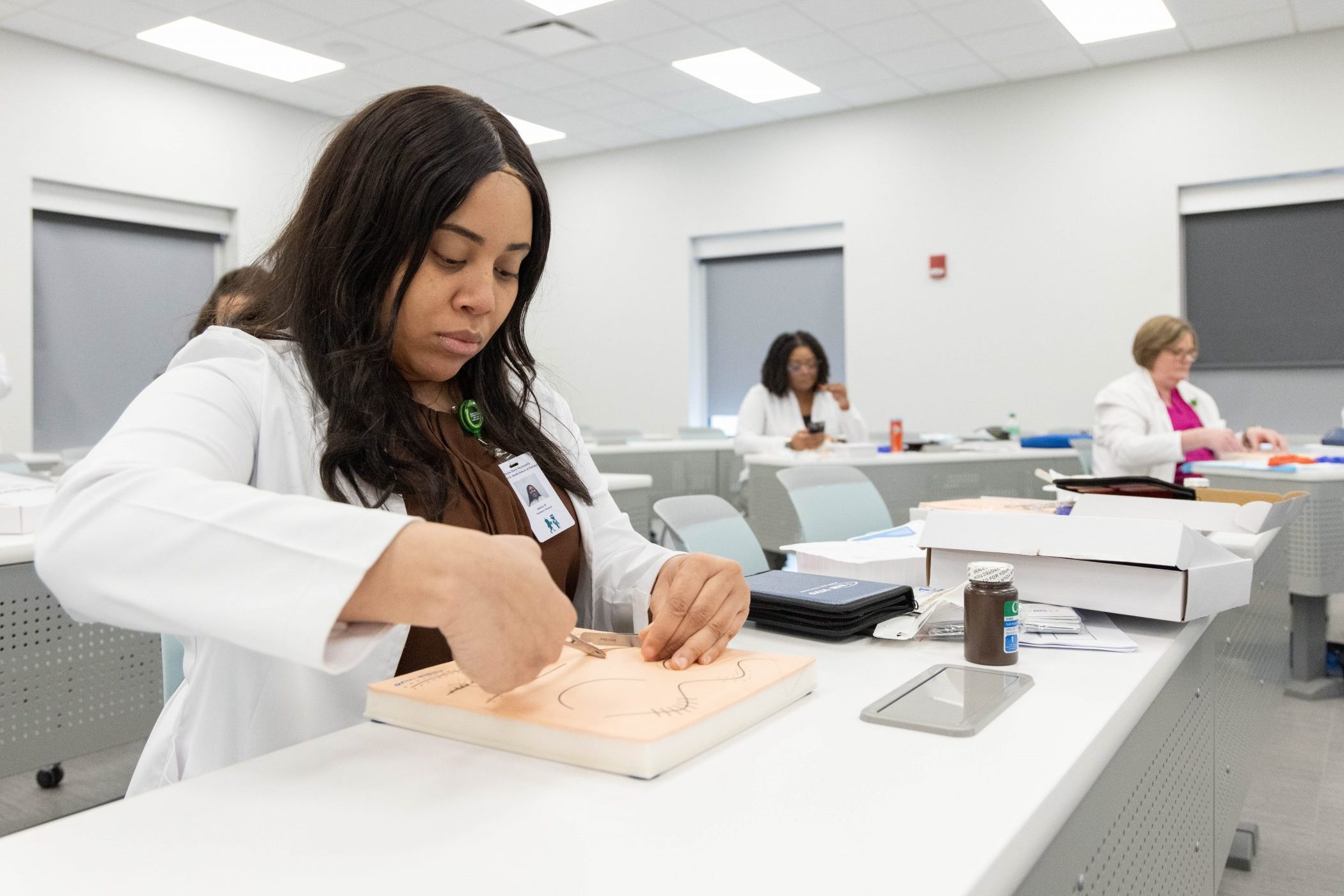 Woman in a lab coat practicing suturing on training pad in a classroom, with others working in the background.