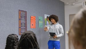 Art entrepreneurship student presenting a design project to peers in class, with creative posters displayed on the wall behind them.