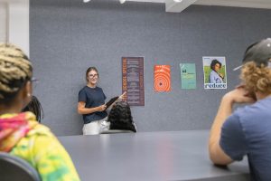 Design student presenting a project to peers in a classroom, with creative posters displayed on the wall.