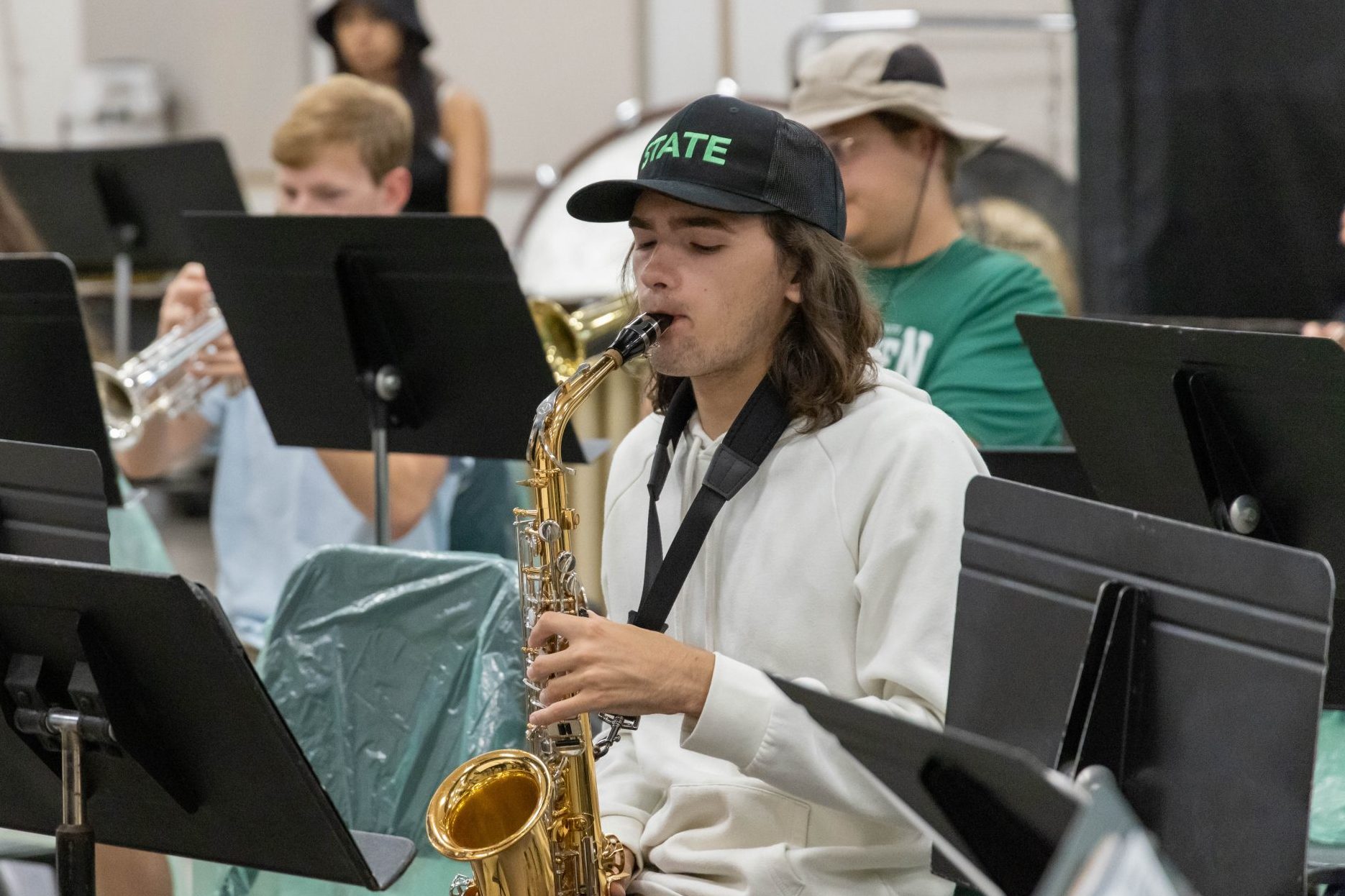 University band student playing saxophone in rehearsal, surrounded by classmates performing on trumpets and percussion.