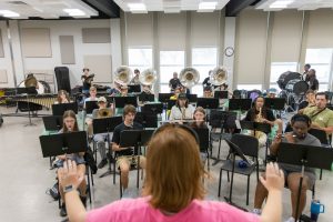 University band students rehearsing under the direction of a conductor, with woodwinds, brass, and percussion instruments filling the classroom.