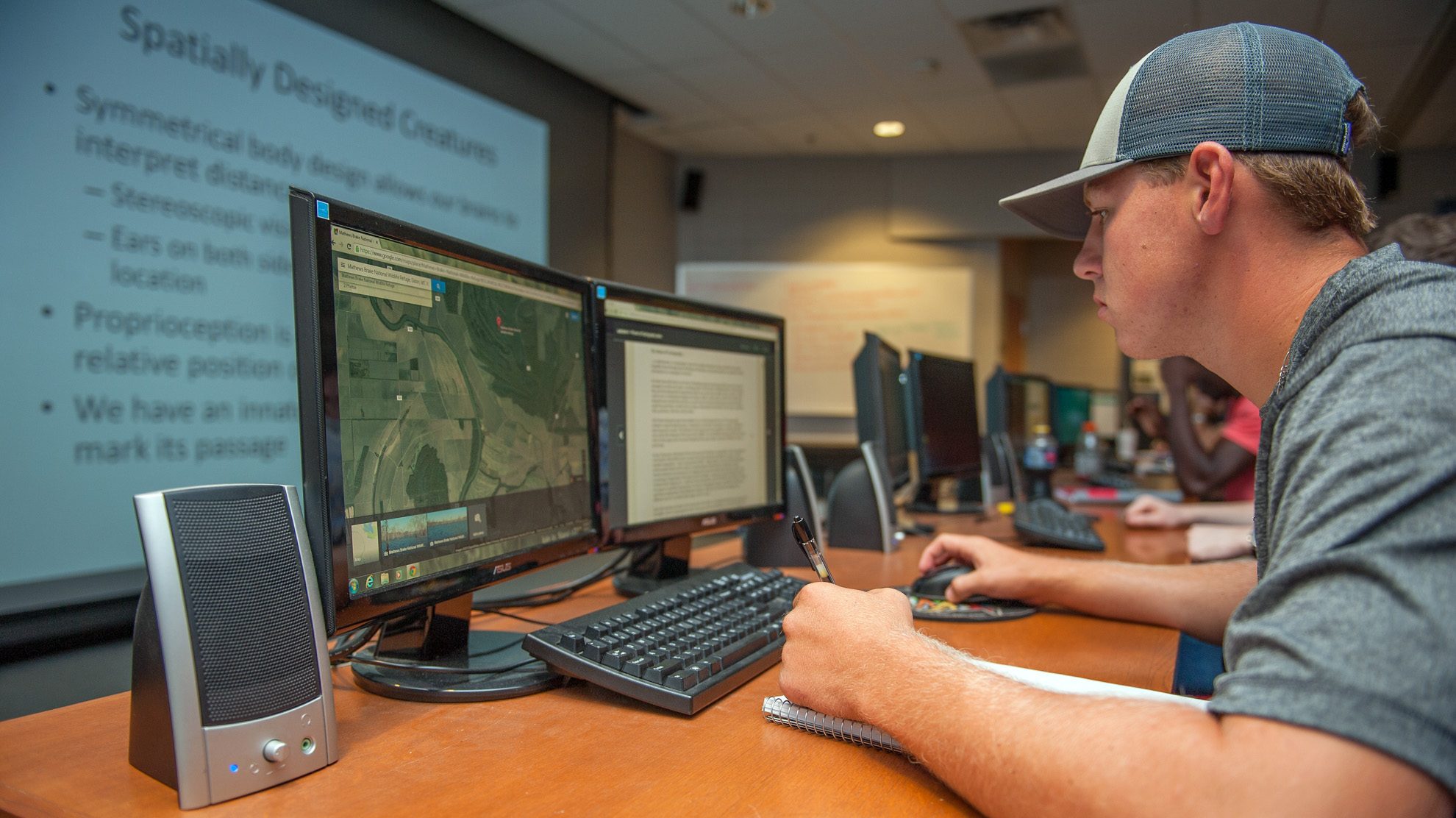 Student analyzing satellite maps on dual monitors in a geospatial information technologies lab.