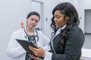 Two women talk in a professional setting; one in a lab coat and stethoscope, the other holding a tablet and dressed in business attire, discussing something work-related.