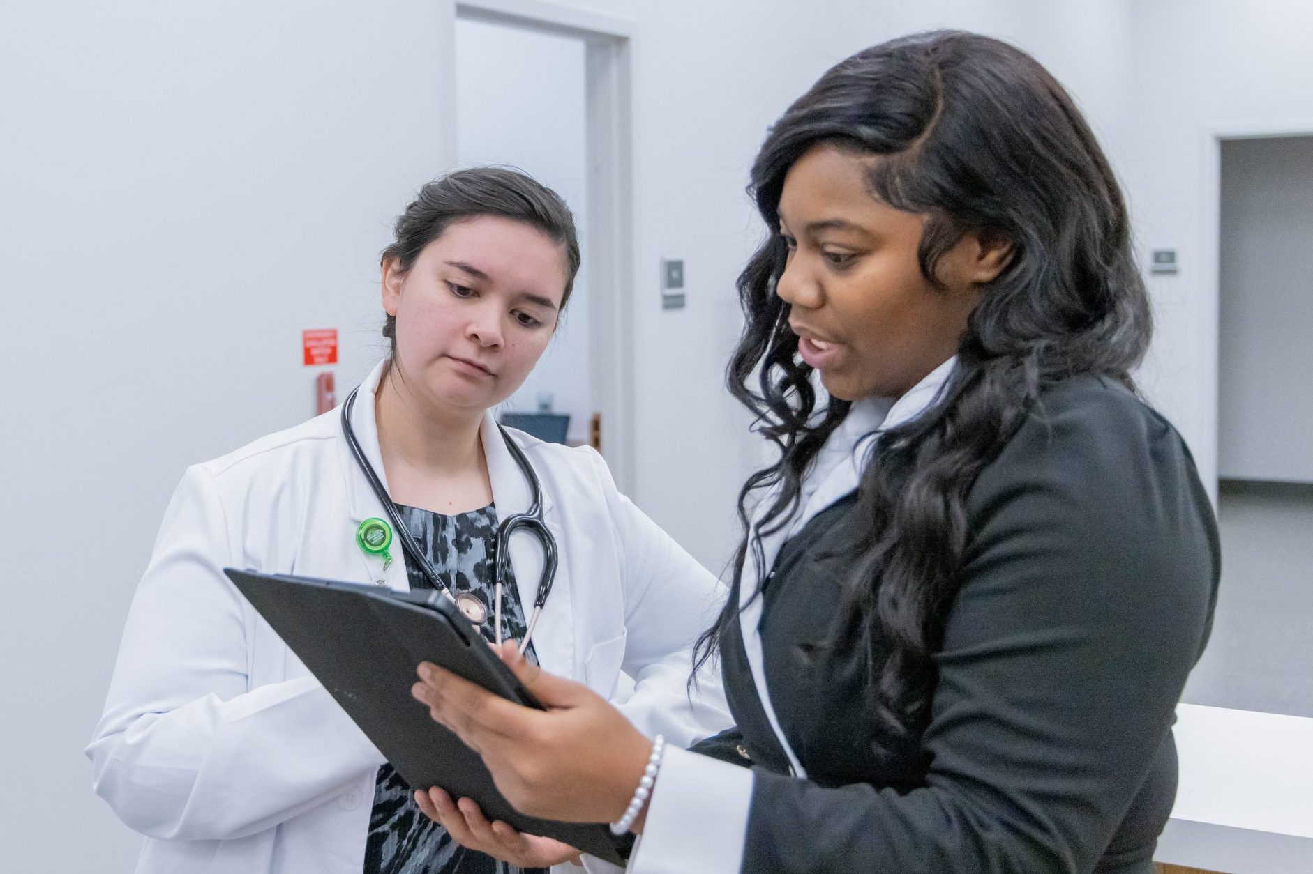Two women talk in a professional setting; one in a lab coat and stethoscope, the other holding a tablet and dressed in business attire, discussing something work-related.