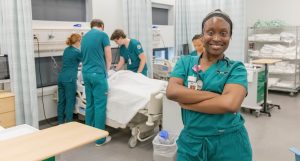 Nursing student standing in a clinical simulation lab while other students provide patient care training.