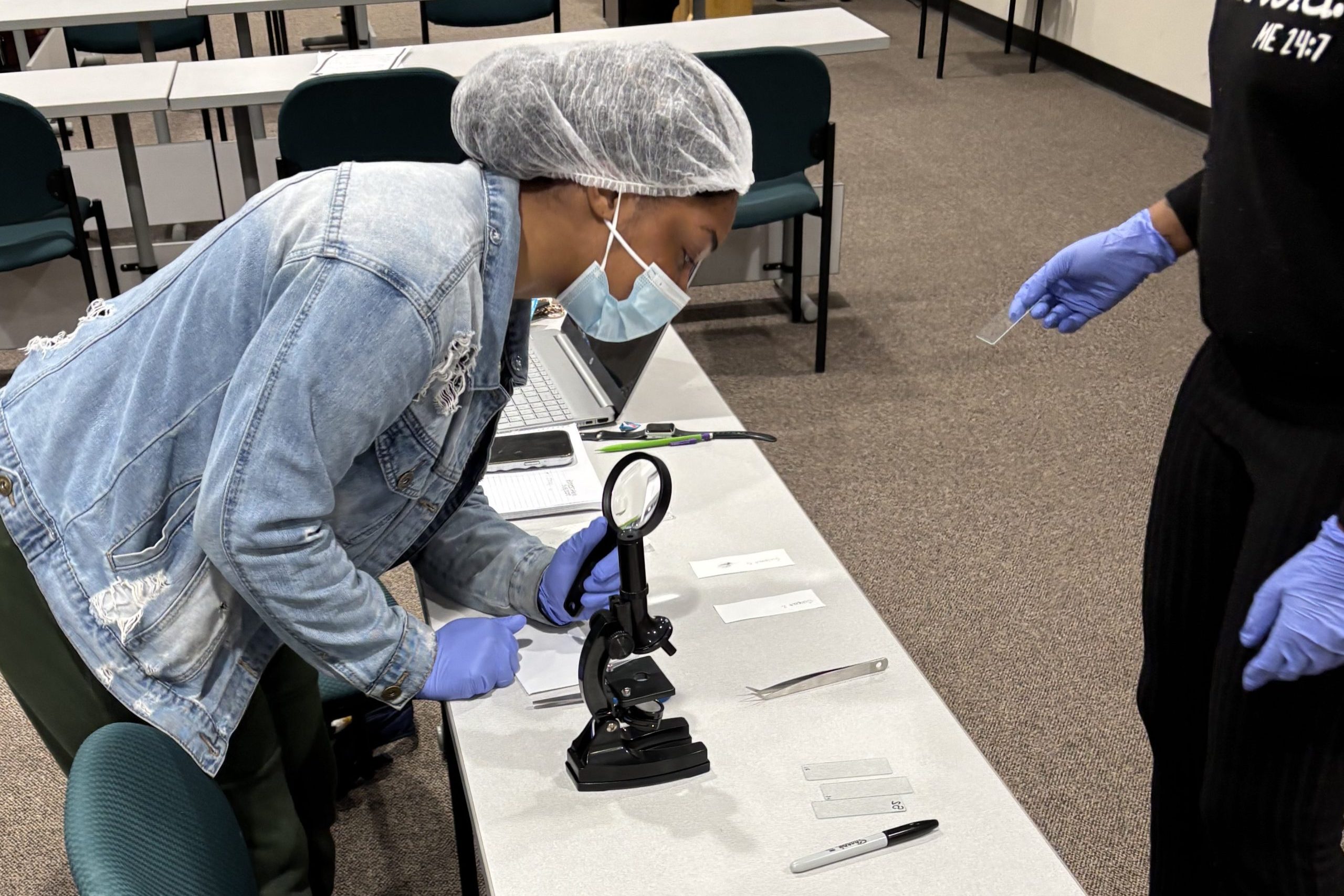 A student wearing gloves, a face mask, and a hair covering examines evidence with a magnifying glass at a table in a forensic science classroom, while another individual with gloves observes nearby.