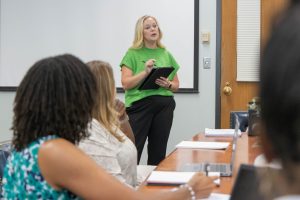 A woman in a green shirt stands at the front of a classroom holding a tablet and speaking, while several students sit at a table facing her with notebooks and materials.
