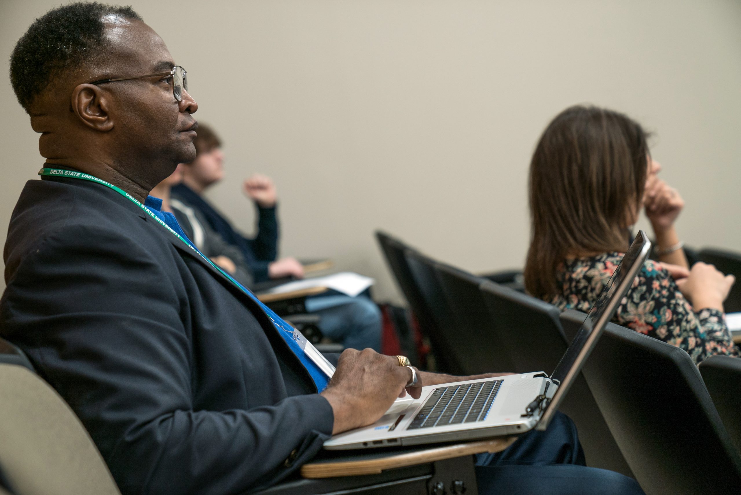 A man in a blazer attentively listens during a lecture or presentation, seated with a laptop on his lap in a lecture hall with other attendees nearby.