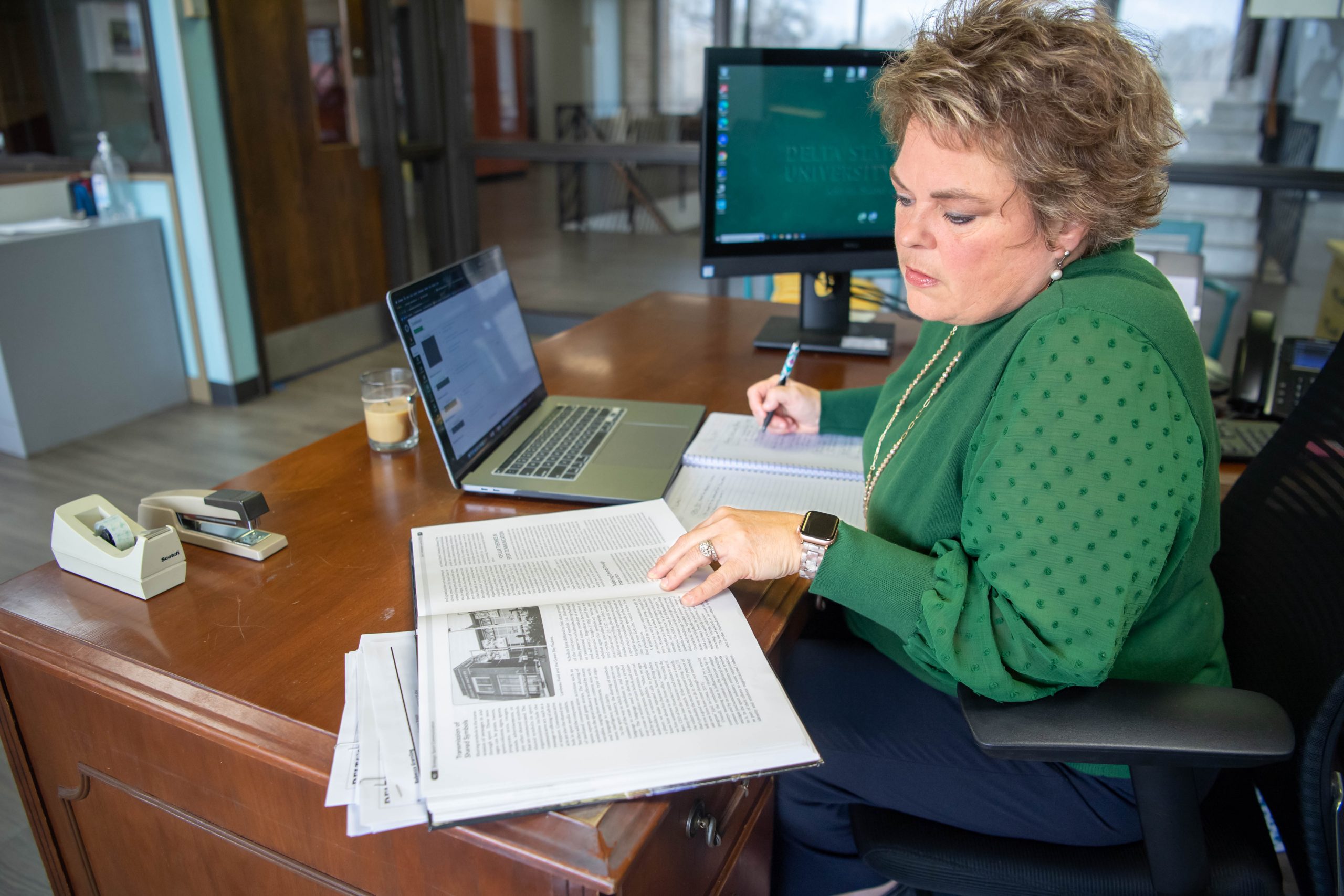 A woman in a green sweater reviewing documents and taking notes at her desk with a laptop and monitor.