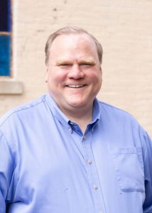 Photograph of white man in blue shirt in front of whitewashed brick wall with blue stained glass windows.