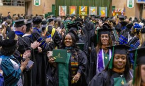 Delta State graduates in caps and gowns walking through a commencement ceremony, smiling and holding diplomas as faculty applaud.